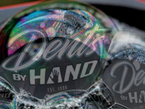 a close up image of some soap bubbles with an iridescent shimmer at the tops of the bubbles. in the reflections of the bubbles are some superimposed images of the Dents By Hand logo to make it appear as if they are a reflection.